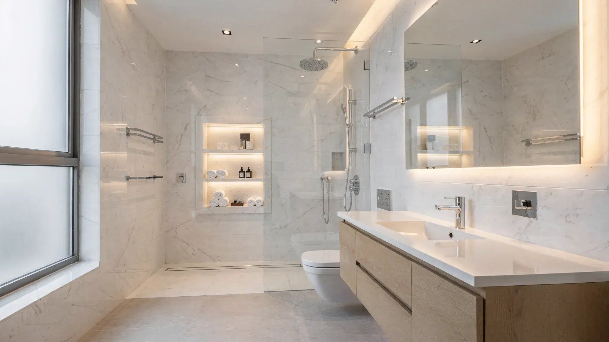 A bright modern bathroom in Dubai with marble-look wall tiles, a floating vanity, warm lighting, glass shower partition, chrome fixtures, and neatly integrated storage.