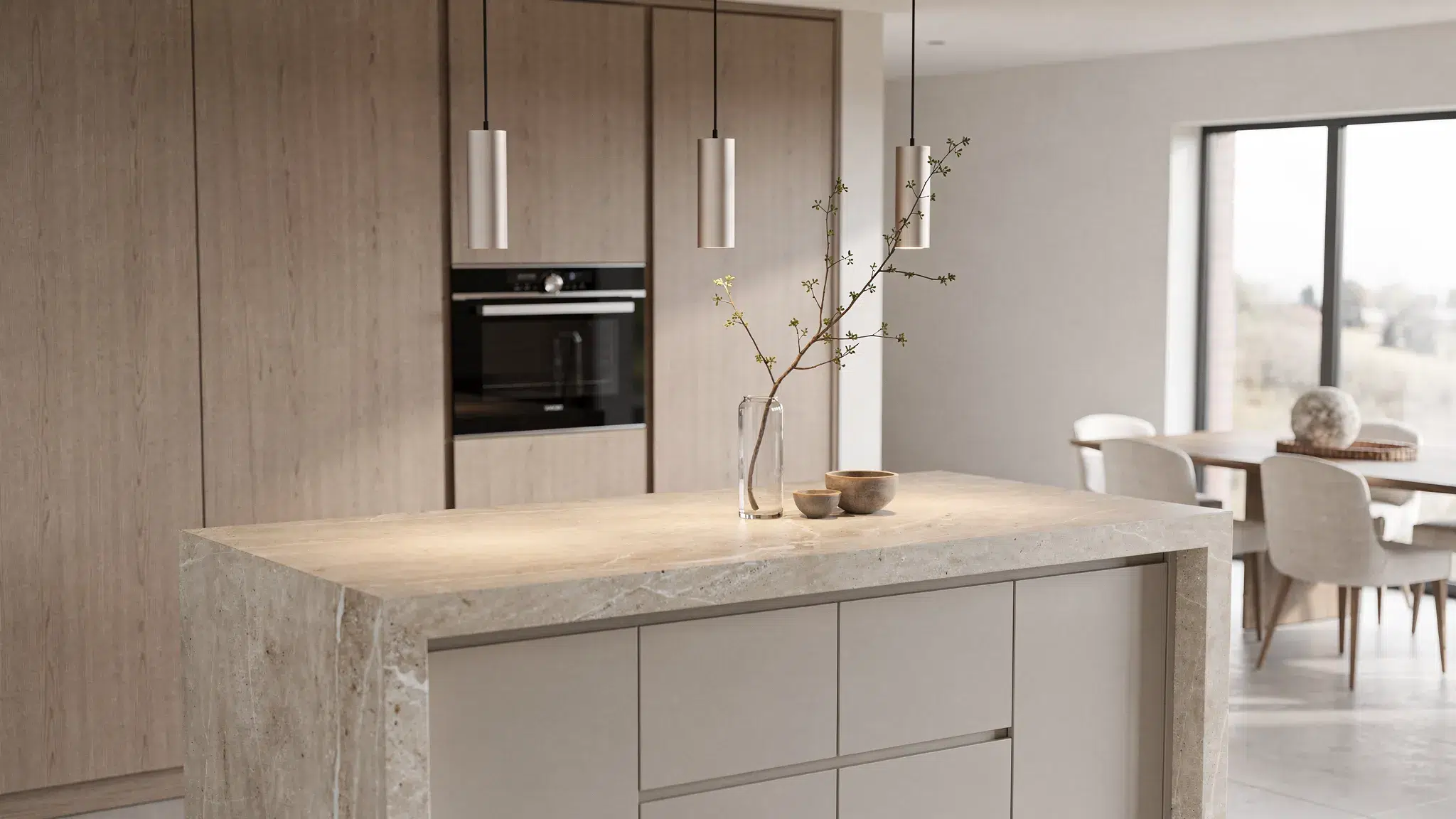 Medium close-up view of a large stone kitchen island with integrated concealed storage and warm wood cabinetry behind it; pendant lights hang above and a refined dining area is visible beyond, all in a calm neutral palette.