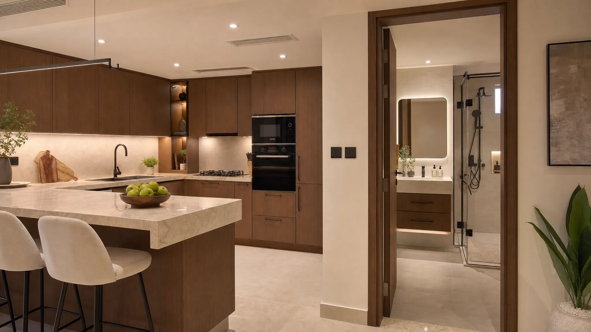 Medium-wide view of a renovated Dubai kitchen connected to a bathroom through an open doorway, showing warm cabinetry, stone countertops, layered lighting, a floating vanity, and a glass shower with neutral tiles.